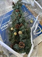 Close-up photo of small artificial holiday tree decorated with pinecones and golden balls, inside a container with plastic wrapping.