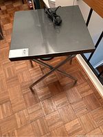 Top angled view of the metal table stand showing the square grey tabletop surface and the attached power cord coiled on top.