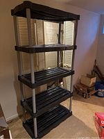 Front view of the 5-tier shelving unit showing the black plastic slatted shelves and silver metal support rods standing in a basement area.