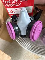 Close-up of 3M dust and mist respirator with purple/pink changeable cartridges attached, gray body, black edges.