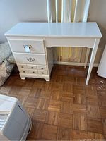 White wooden student desk with three drawers, showing front and side profile, on parquet floor with natural light from window
