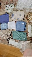 Multiple folded tablecloths and linens laid out on bed: includes plain blue, green, beige and light blue, blue/white embroidery, floral prints