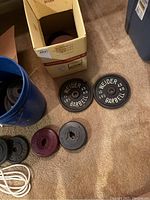 Various metal and heavy plastic weight plates, some stacked inside a cardboard box, adjacent to a blue plastic bucket and white jump rope coiled on the floor.