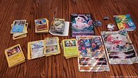Displays multiple small stacks of Pokémon cards plus 3 large oversized cards and a Pokémon coin on wood surface.