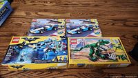 Overview of all four Lego sets in unopened boxes on wooden floor, including two Creator Race Car sets, one Batman Mr. Freeze set, and one Creator T-Rex Dinosaur set.