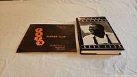 Photo showing Isy's Supper Club menu closed with the cover design and Oscar Peterson biography book side by side on a light surface.