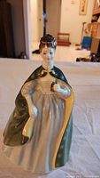 Front view of a Royal Doulton porcelain figurine showing a woman with elaborate hairstyle, green cloak with floral details, and cream dress, holding an object in her right hand.