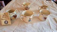 Five Royal Doulton Bunnykins mugs arranged on a white cloth with a Royal Doulton Bunnykins collection booklet beside them, showing various Bunnykins character artwork.