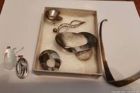 Photo showing the jewellery laid out: two pairs of earrings, two brooches in a box, and a curved necklace band on the side. Silver items show tarnish.