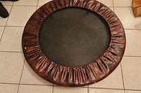 Top view of small round trampoline on tiled floor showing brown padded edge and black jumping mat