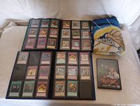 Two Cardguard folios open showing various Magic: The Gathering and Yu-Gi-Oh trading cards inside, with a folded Yu-Gi-Oh blanket beside them and Yu-Gi-Oh DVD case laying flat
