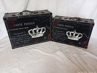 Front view of two decorative trunks, larger and smaller, both black with white crown and postcard script graphics