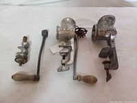 Two vintage manual meat grinders shown side by side on a cloth surface. Both grinders have metal bodies, wooden handles, and clamp bases. One grinder shows the auger separate and the other has the crank attached.