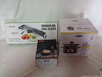 Three boxed kitchen items on white cloth background: a mandoline dial slicer, a slow cooker, and a skull mini waffle maker.