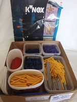 Photo showing multiple containers filled with K'NEX rods and connectors of various colors including red, yellow, gray, dark blue, and purple. The K'NEX instruction booklet is placed behind the containers.