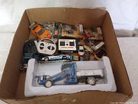 Box with assortment of toy cars and vehicles including blue tractor flatbed model, orange sports car, remote control, toy motorcycles in packaging