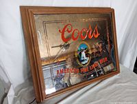 Side angled view of the Coors lighted bar sign showing the wooden frame, reflective/mirrored surface, red Coors logo, and visible chip damage on the upper right corner of the frame.
