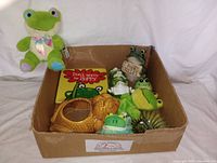 Photo of the entire lot showing a cardboard box holding assorted ceramic and plush frogs, and a frog sign.