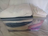 Clear plastic storage bin filled with folded assorted towels showing white and beige towels on top, with blue, pink, and patterned towels inside the bin.