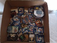 Overview photo showing a cardboard box filled with assorted trading cards featuring Pokémon, Magic The Gathering, and Yu-Gi-Oh cards along with two collectible Pokémon card tins.