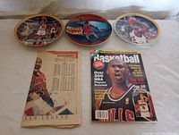 Three round collectable plates featuring Michael Jordan in Chicago Bulls uniform in basketball action scenes plus a basketball yearbook magazine and a newspaper with Michael Jordan coverage.