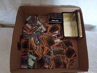 Wide view of cardboard box with assorted trading cards scattered inside including Yu-Gi-Oh cards and some sleeved cards, along with two metal tins for storage.