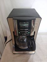 Front view of Mr. Coffee programmable coffee maker showing stainless steel and black plastic construction with glass carafe and black handle on warming plate.