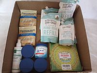 Overview of all items in the lot inside a cardboard box showing bath treatment packets, body cleanser bottle, blue gel jar, refill pouches for hand soap and sanitizer, and incense cones box.