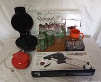 Full lot view showing waffle maker open, boxed Oneida item, Nostalgia My Mini Griddle, glassware, mug and tea lights arranged on table.