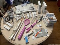 Photo showing two white Nintendo Wii consoles, six Wii remotes (five white, one black), various cables and nunchucks, and multiple Wii accessories including tennis racket, golf club, and gun attachments. Also shows a stack of Wii game discs and several operation manuals.