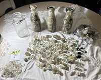 Wide view of three antique glass milk bottles filled with small tumbled stones, a collection of loose polished stones and raw crystals arranged on a lace cloth-covered table.