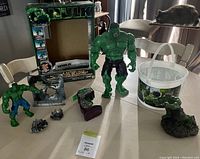 Wide shot of Hulk collectible lot items including two Hulk figures, packaging box, bucket, and accessory stands on table