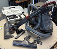 Photo showing Dyson canister vacuum with hose and various attachments including motorized and brush tools spread on table with original box open in background