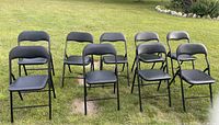 Nine black folding chairs arranged on grass outdoors, showing front and side views, displaying metal frame and padded vinyl seats and backs.