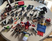 Overview of various action figures and toys on a round table surface, displaying figures in both standing and lying down positions with assorted accessories and small vehicles.