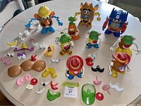 Multiple Mr. & Mrs. Potato Head figures and parts arranged on table, including two dressed as Transformers characters Optimus Prime and Bumblebee, and one with a construction hat and blue pigtails. Various interchangeable parts laid out around the figures.