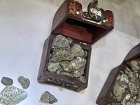 Photo showing a small wooden treasure chest filled with raw pyrite nuggets and some pyrite pieces placed next to it.