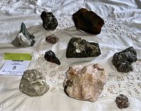 Collection of raw mineral and gemstone rocks placed on lace tablecloth including agate slice, quartz clusters, and various rock specimens.