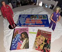 Photo showing Six Million Dollar Man and Bionic Woman action figures, Six Million Dollar Man board game box, and two Bionic Woman comic books on a white lace tablecloth.
