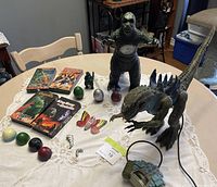 Full view of vintage Godzilla action figures, VHS tapes, collectible eggs, butterfly figure, small figurines, and handheld toy on table.