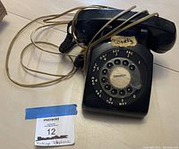 Photo showing the black rotary phone with visible rotary dial, handset, and cord. 'Lookers' label is visible on the front.