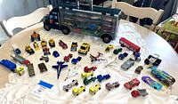 Top view of the entire toy vehicle lot spread on a white lace tablecloth, showing the large transport truck, multiple small vehicles including cars, trucks, planes, and motorcycles.