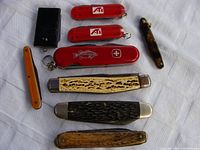 Top-down view of nine assorted pocket knockdown cutters arranged on white cloth, showing various handle materials including plastic, bone, and metal. Condition ranges from lightly worn to missing parts.