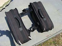 Two black Joe Rocket motorcycle saddle bags connected by straps and lying flat on a concrete surface in sunlight.