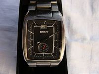 Close-up view of DKNY wristwatch showing rectangular black dial, silver markers, date window, and small seconds subdial. Visible light scuffs on stainless steel case.