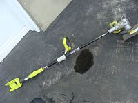 Full length photo of Ryobi cordless string trimmer showing shaft, handle, and cutting head on a black concrete surface.