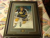 Full view of framed Bobby Orr print showing player in Bruins uniform, signed 'A' on jersey, on quilt background.