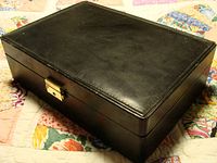 Closed black watch box showing textured surface with visible depression marks on the top and a gold lock centered on the front.