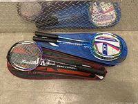 Three badminton rackets laying flat, each with a cover; two blue covers and one black with red accents.