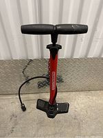 Full view of red Supercoach floor bike pump with black handle and hose, placed in front of metal diamond plate wall.